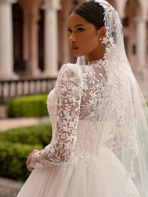 Νυφικό με μανίκια και μπούστο από ιδιαίτερη δαντέλα με πέπλο εξτρά.